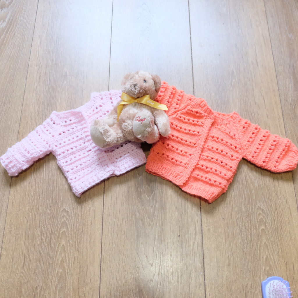 Examples of the cardigan, knitted in orange and crocheted in pink, lying on the floor with a small teddy bear for scale.